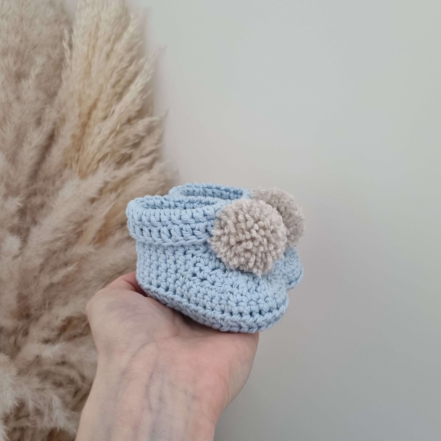 A hand cradles a pair of blue crocheted baby bootie, illustrating the warmth and care in its handmade crochet design.