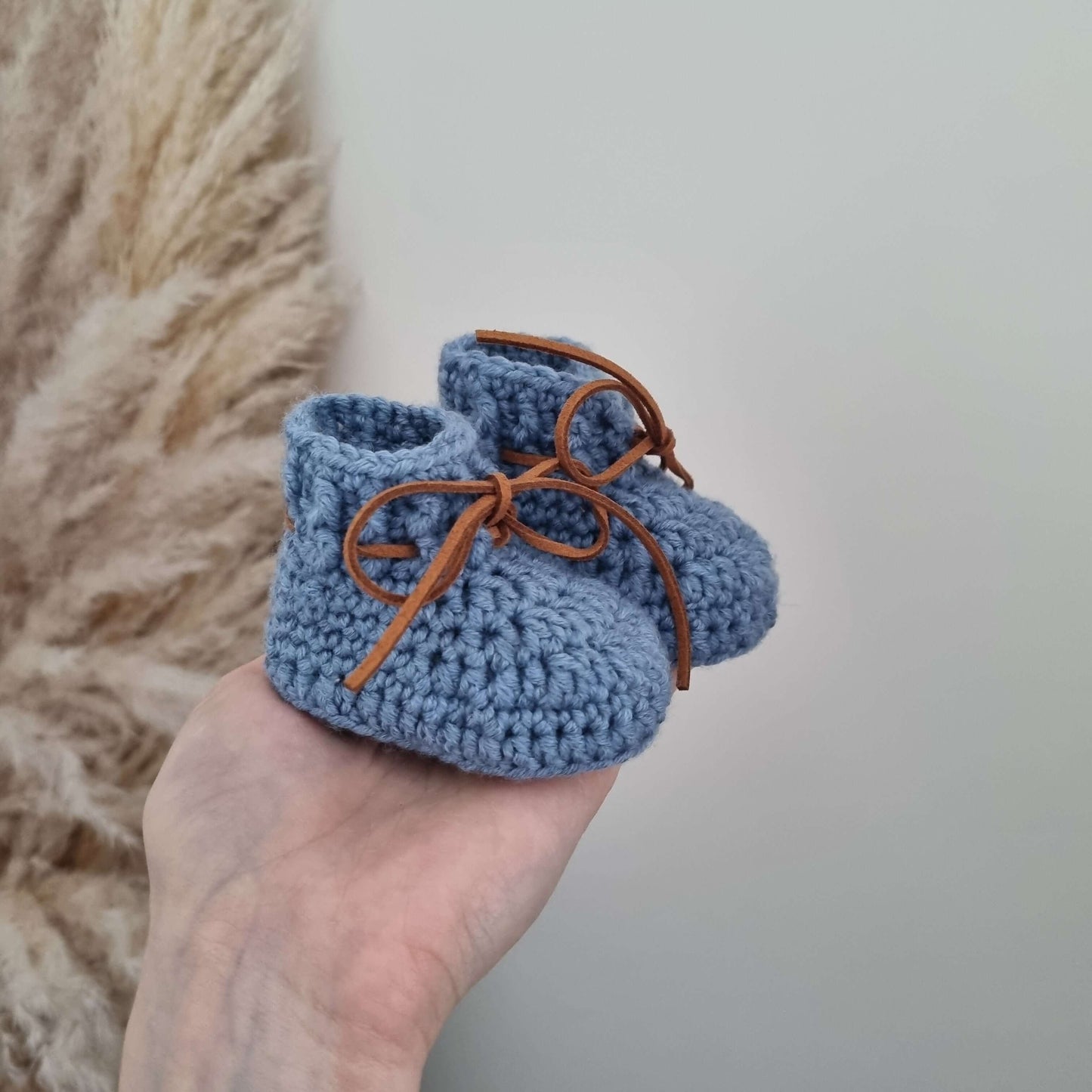 A hand gently holds a pair of blue crocheted baby booties made from soft Merino wool.