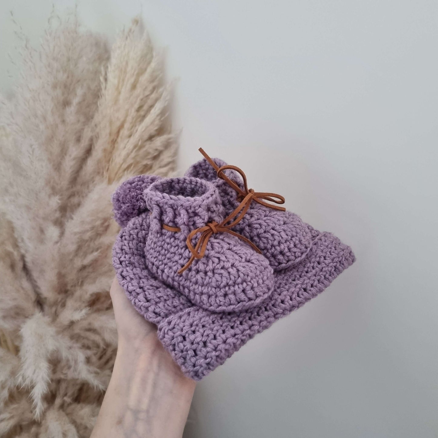 A hand presents a lovely purple baby hat with a matching pair of baby booties, beautifully crocheted from Merino wool.