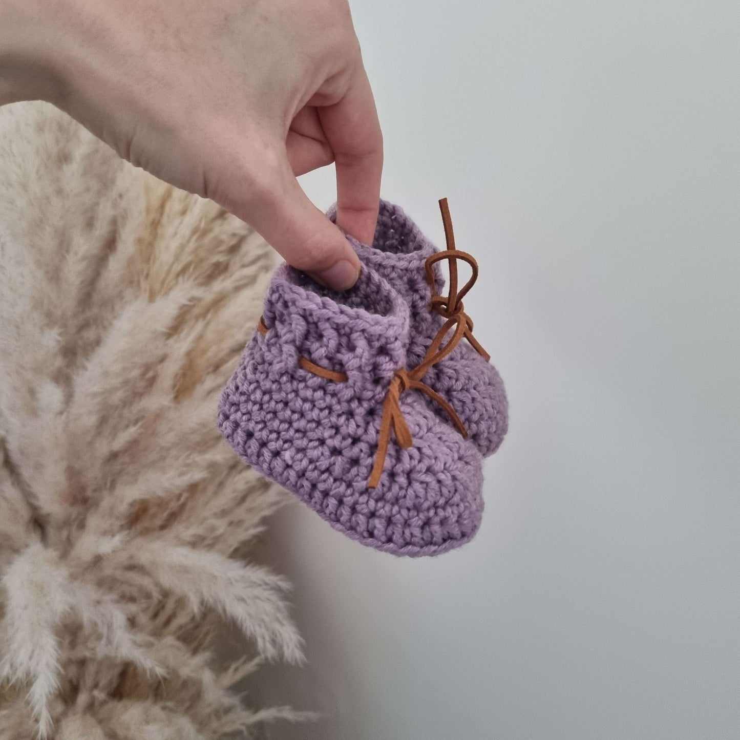 A hand gently holds a pair of purple crocheted baby booties made from soft Merino wool.
