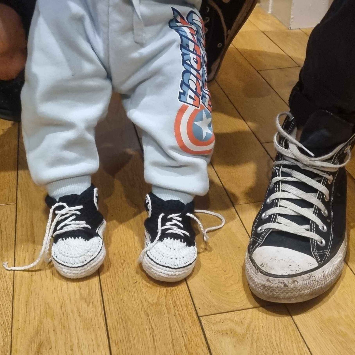A baby in cute black crochet Converse sneakers stands next to a man in stylish matching sneakers, both sharing a sweet moment.
