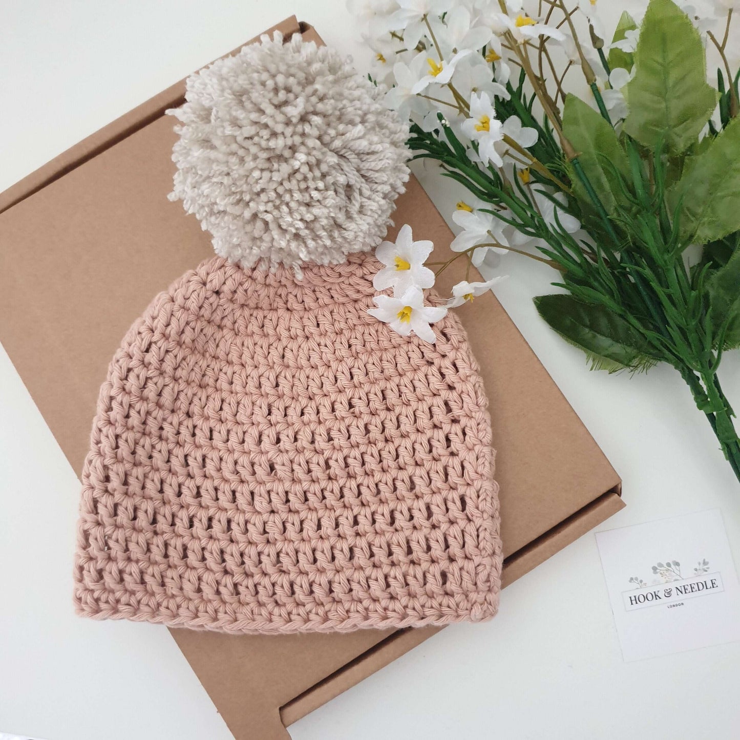 Baby Hat & Boots Set - Clay Pom Pom with soft pom pom detail nestled on a brown box, surrounded by flowers.