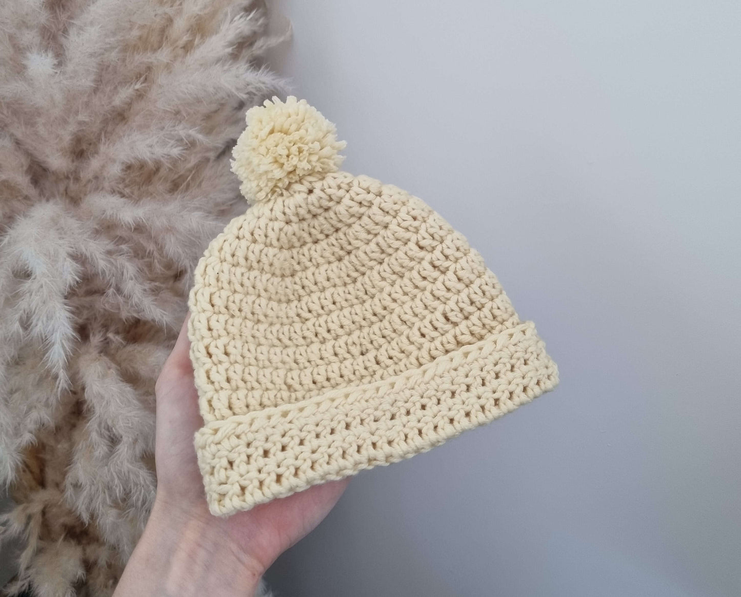 Merino wool baby hat and boots set in custard color with pom-pom on top, held against a soft background.