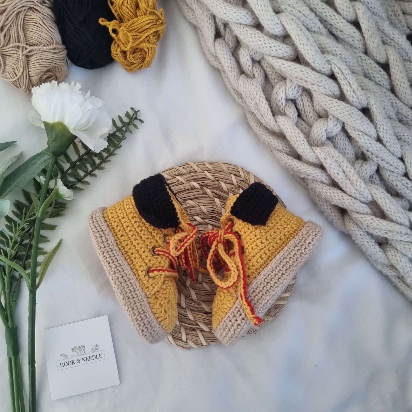 Crochet baby work boots in yellow and black with a lace-up design, hand-crocheted and styled on a woven mat.