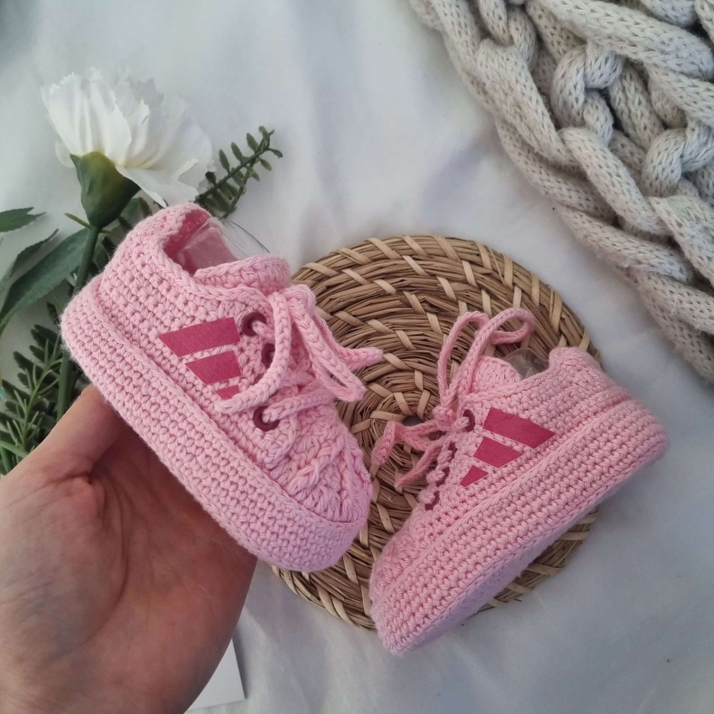 Pink Crochet Baby Sneakers held in hand on a woven mat with greenery, showcasing their handmade design.