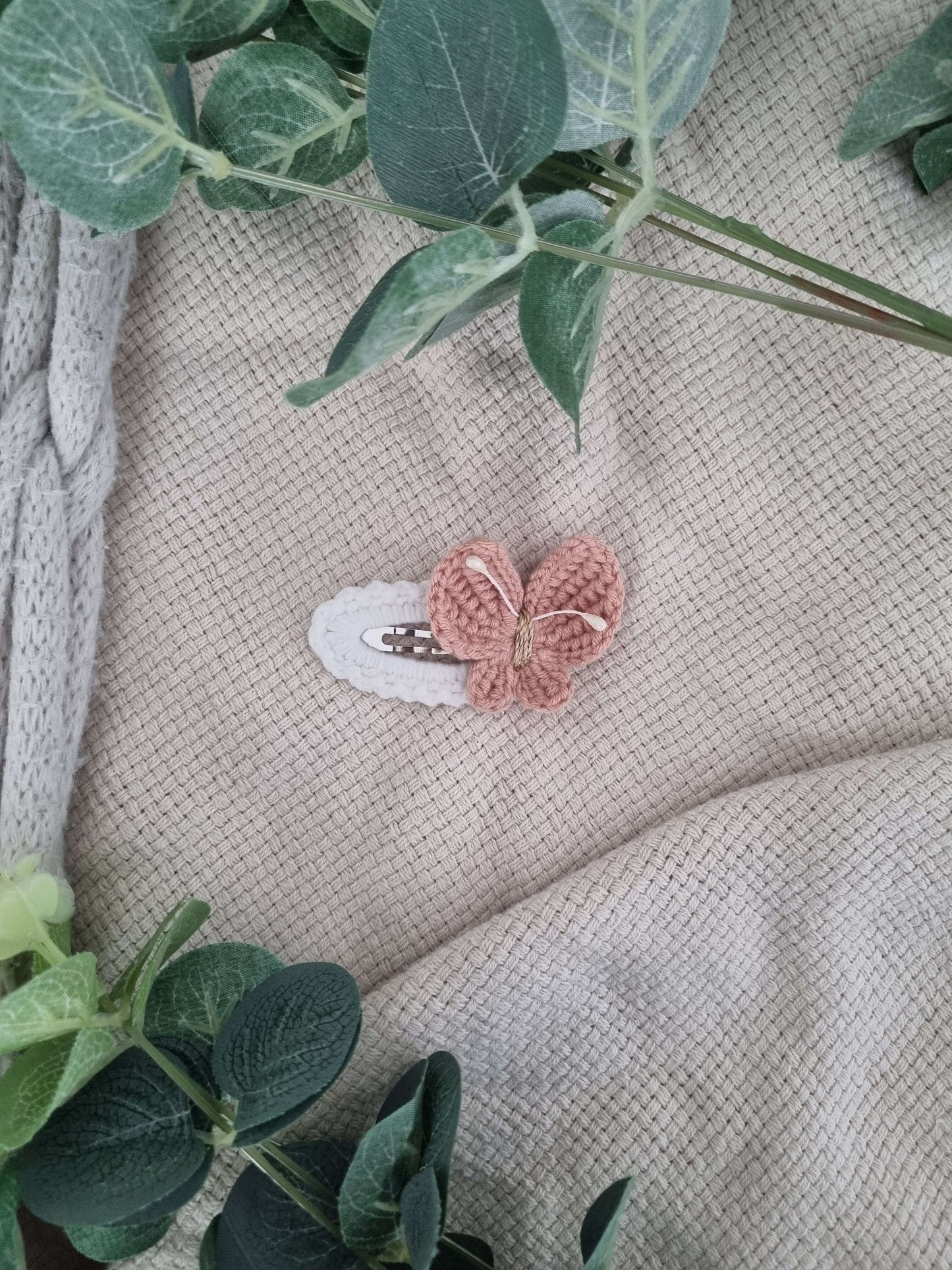 Handmade crochet butterfly hair clip resting on a neutral fabric background surrounded by green leaves.
