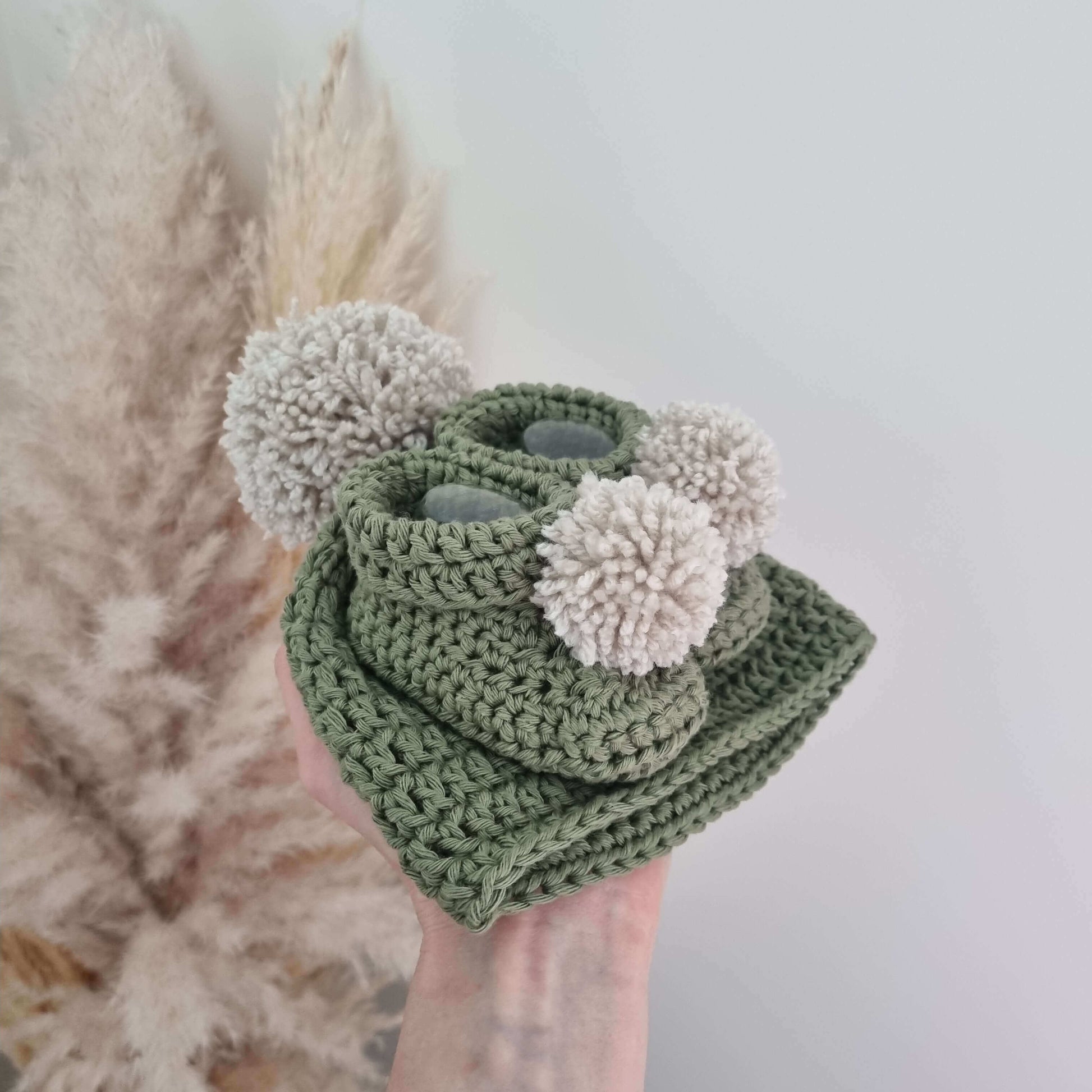 A hand holds a green crocheted baby hat and matching pom pom boots, showcasing a cozy winter accessory set.