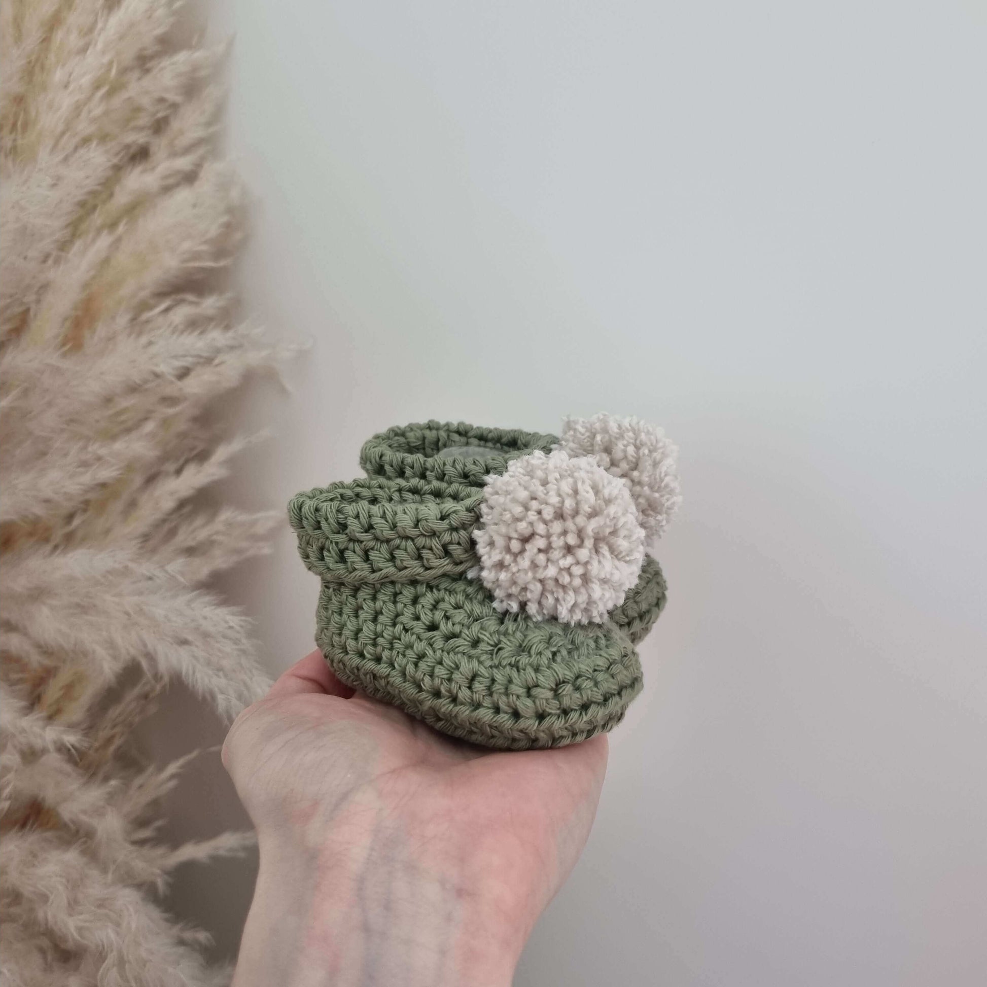 A hand gently holds a pair of green crocheted baby booties, showcasing their delicate craftsmanship and soft texture.