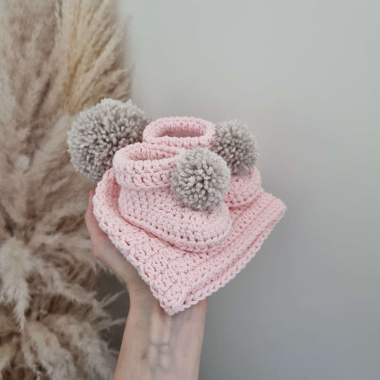 A hand displays a pink crocheted hat adorned with pom poms booties, perfect for a baby, highlighting its charming craftsmanship.