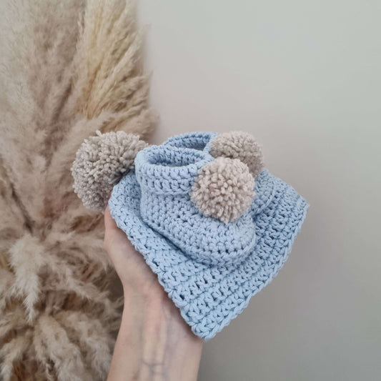 A hand holds a baby blue crocheted hat and matching pom pom boots, showcasing a cozy winter accessory set.