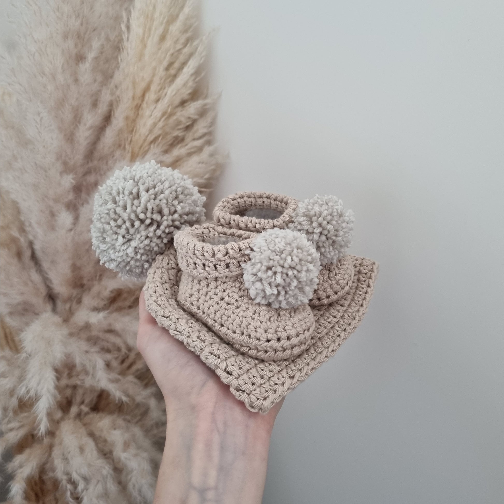 A hand presents a stone coloured crocheted hat with matching pom pom booties, ideal for adding flair to winter outfits.