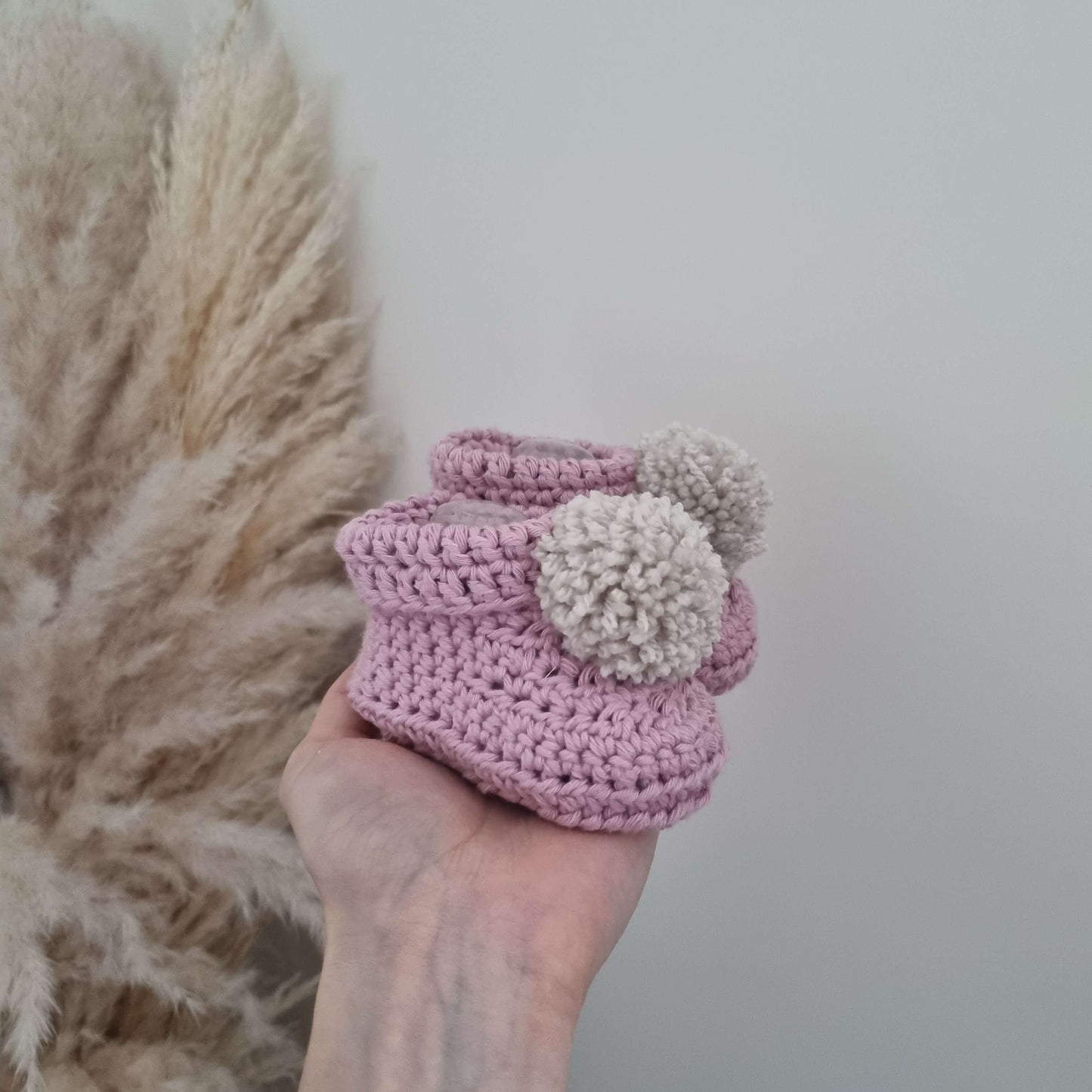 A hand cradles blossom pink crocheted baby booties, illustrating the warmth and care in its handmade crochet design.