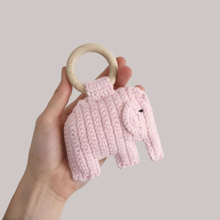 A hand holds a soft, baby pink elephant rattle, showcasing its cute design and vibrant colors.