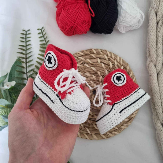 A hand holds a pair of red and white crochet baby Converse sneakers, showcasing their playful design and vibrant colors.