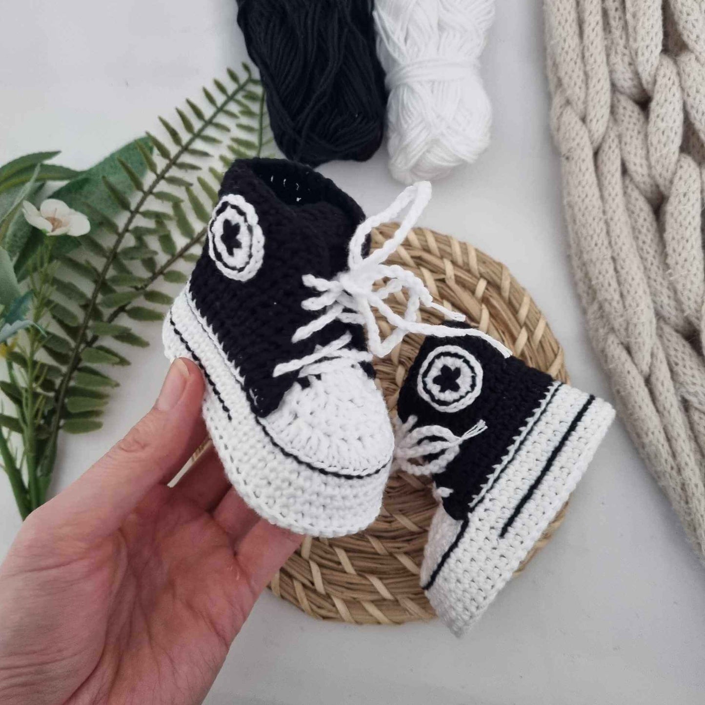 A person holds a pair of black and white crochet baby Converse sneakers, showcasing their adorable design and craftsmanship.