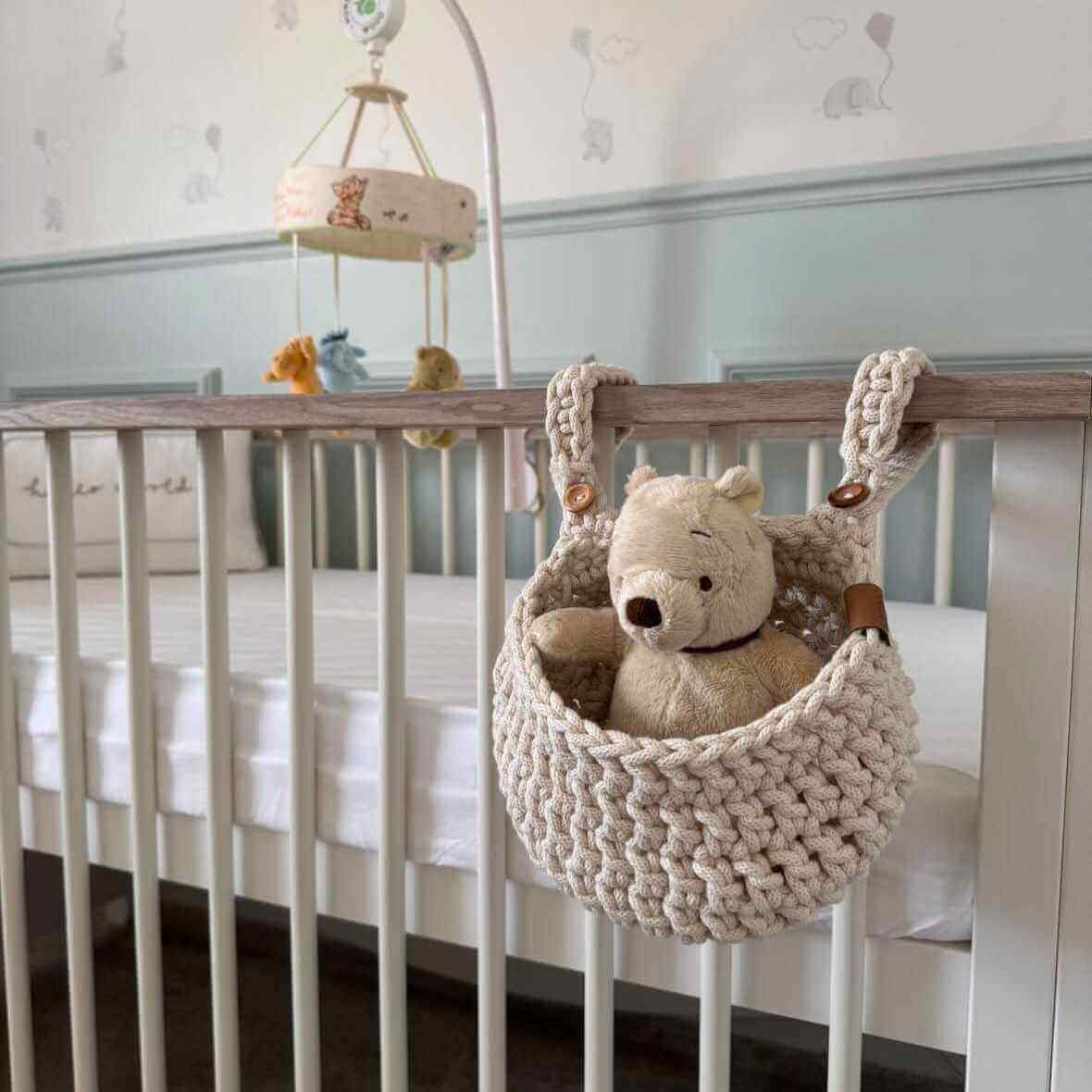 A cute teddy bear hangs from a handmade cot basket in a cozy nursery, adding charm to the baby’s room.