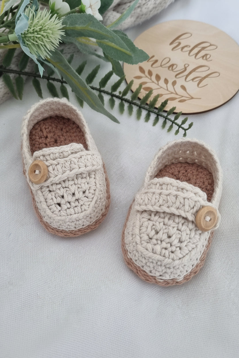 A pair of handmade cream and brown crochet baby booties with wooden buttons, styled on a light fabric background with a wooden sign reading 'hello world' and green foliage.