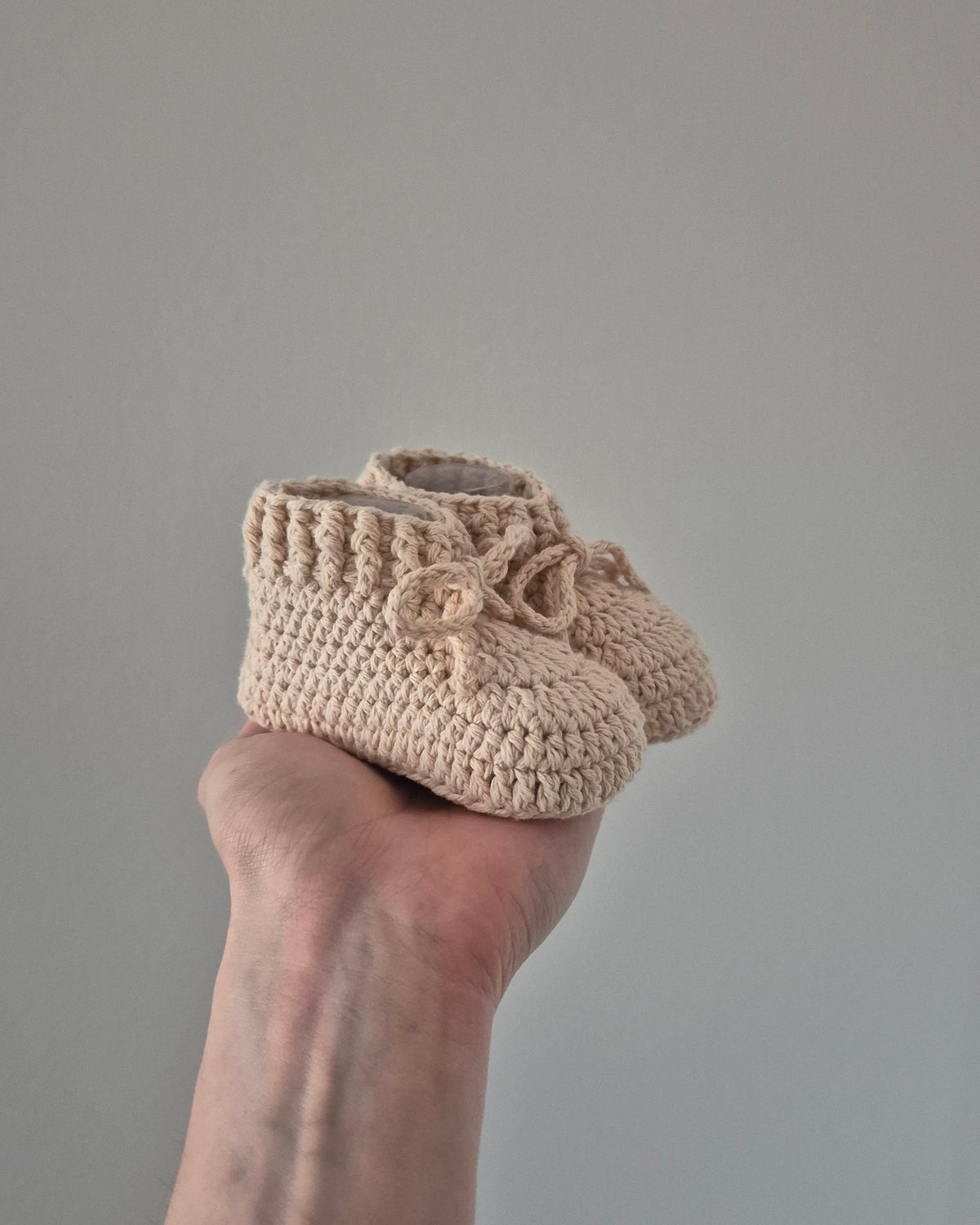 A pair of beige crocheted baby booties with bow details, held in a person's hand against a plain gray background.