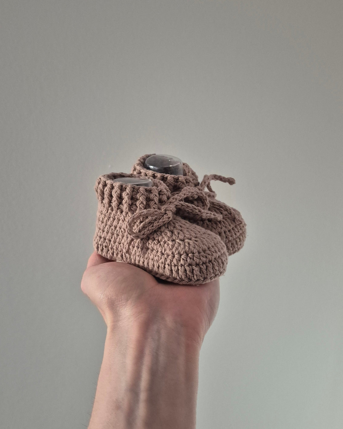 A pair of handmade, chunky crocheted baby booties in a dusty taupe-brown color, held in a person's hand against a plain, light gray background.