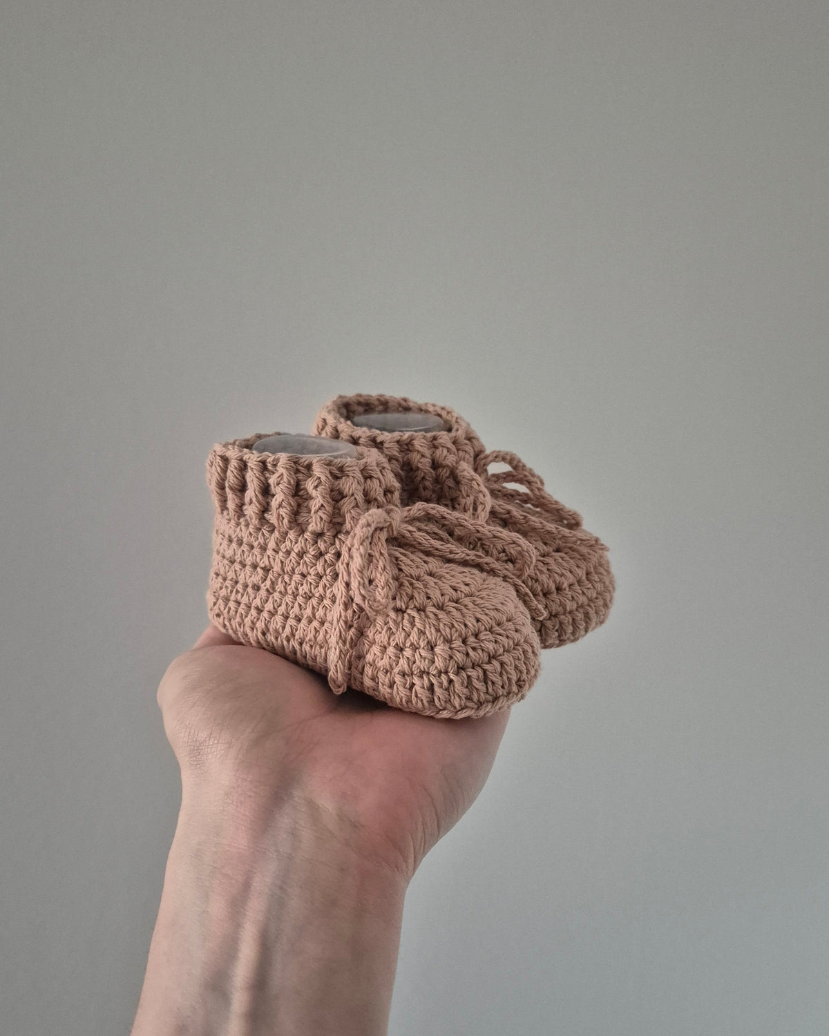 A pair of handmade, chunky crocheted baby booties in a dusty rose pink color, held in a person's hand against a plain gray background.