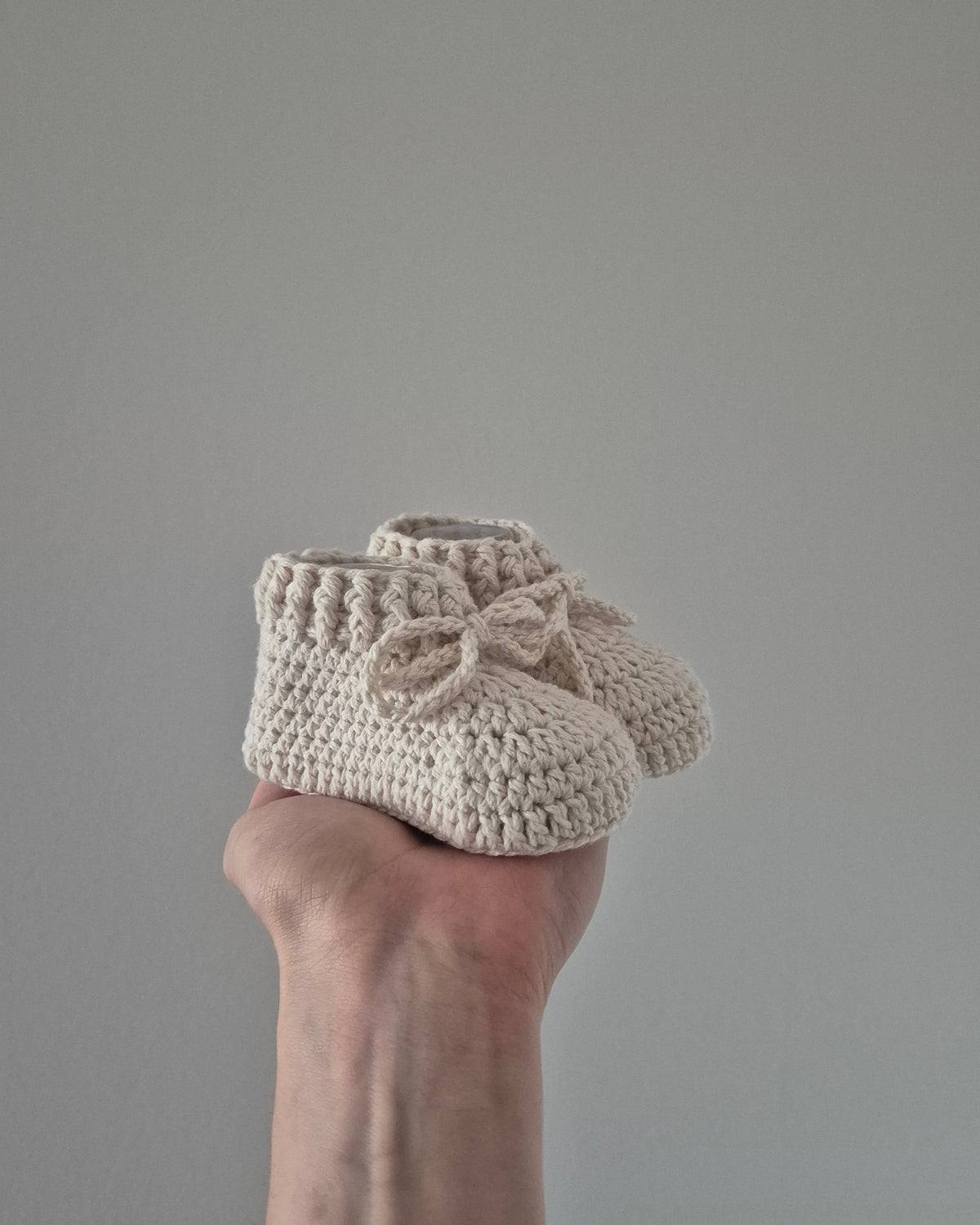 A pair of cream-colored crocheted baby booties with matching bow details, held in a person's hand against a plain gray background.