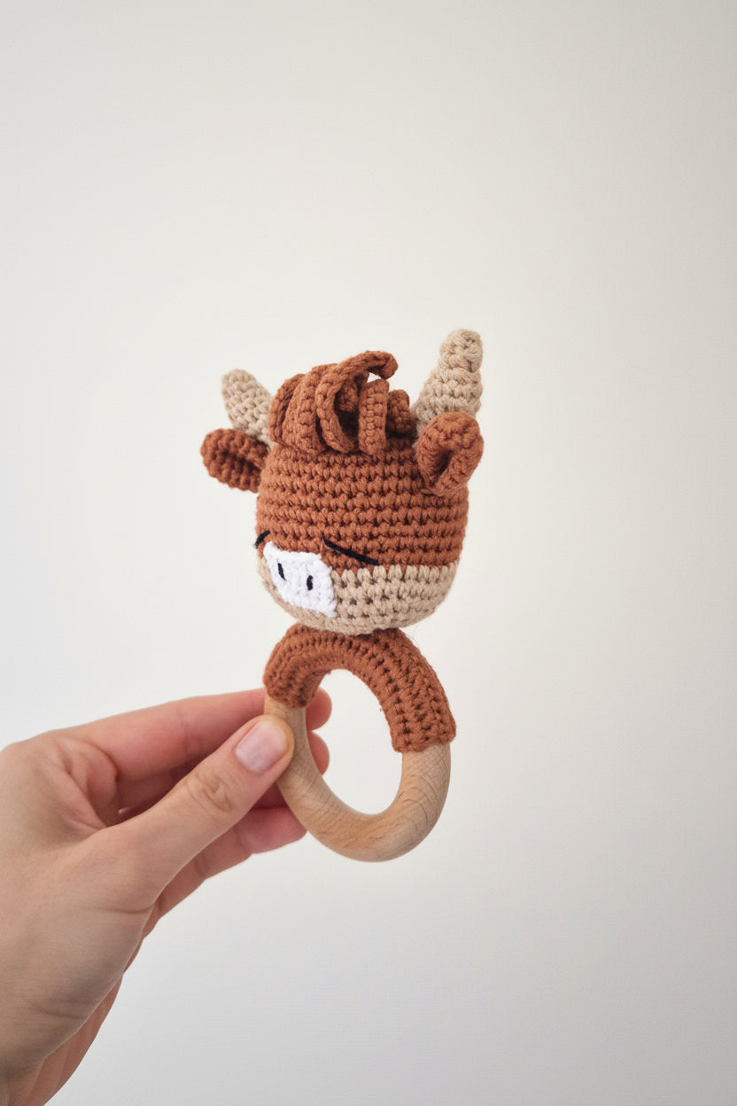 A hand holding a crocheted baby teether rattle shaped like a reindeer head with a wooden ring handle, against a plain light gray background.