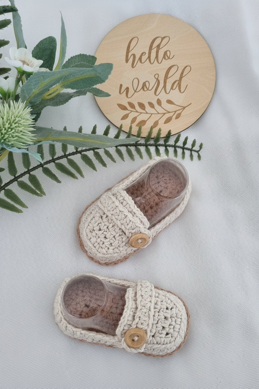 A pair of handmade cream crochet baby booties with wooden buttons, styled on a white textured blanket with a wooden 'hello world' sign and green foliage.