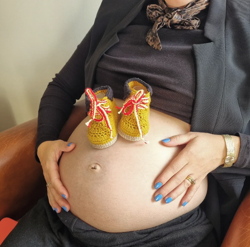 A pregnant mother with blue nail polish rests her hands on her bare belly with a pair of crochet baby sneakers placed on top.