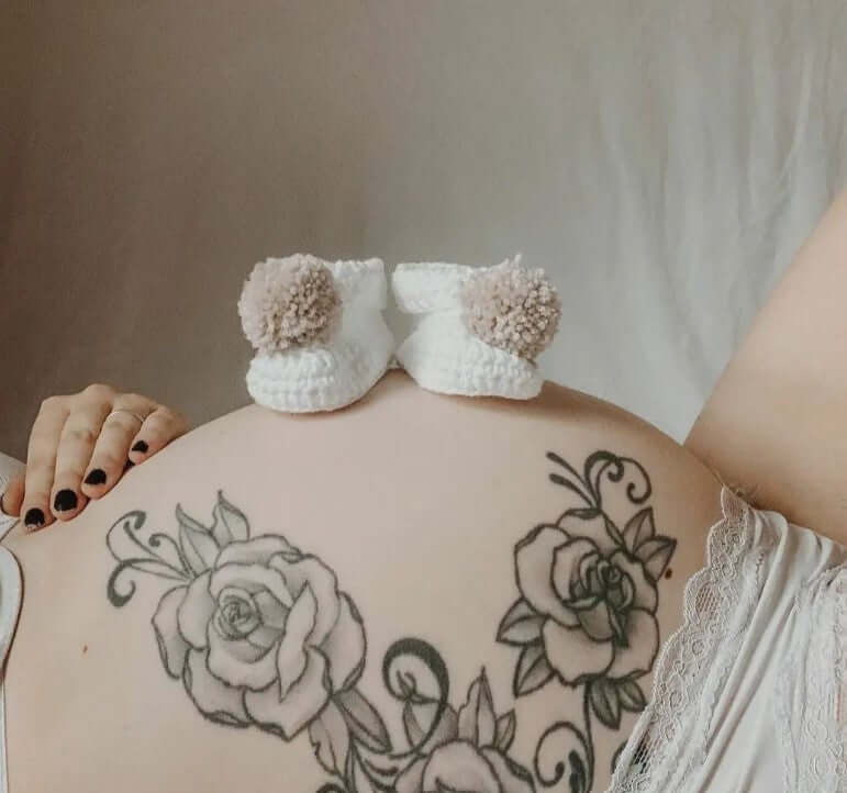 Cotton pom pom baby boots on a pregnant belly with floral tattoo, showcasing playful design and delicate details.