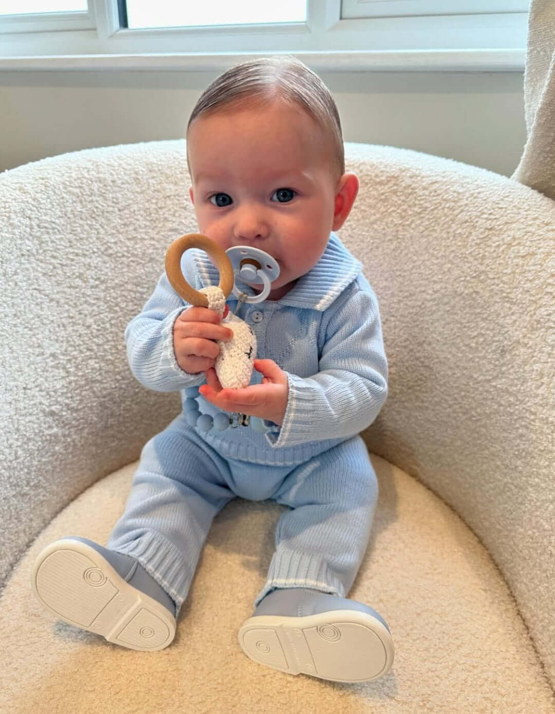 Baby holding a crochet cloud rattle while sitting in a cozy chair, wearing a stylish blue outfit.