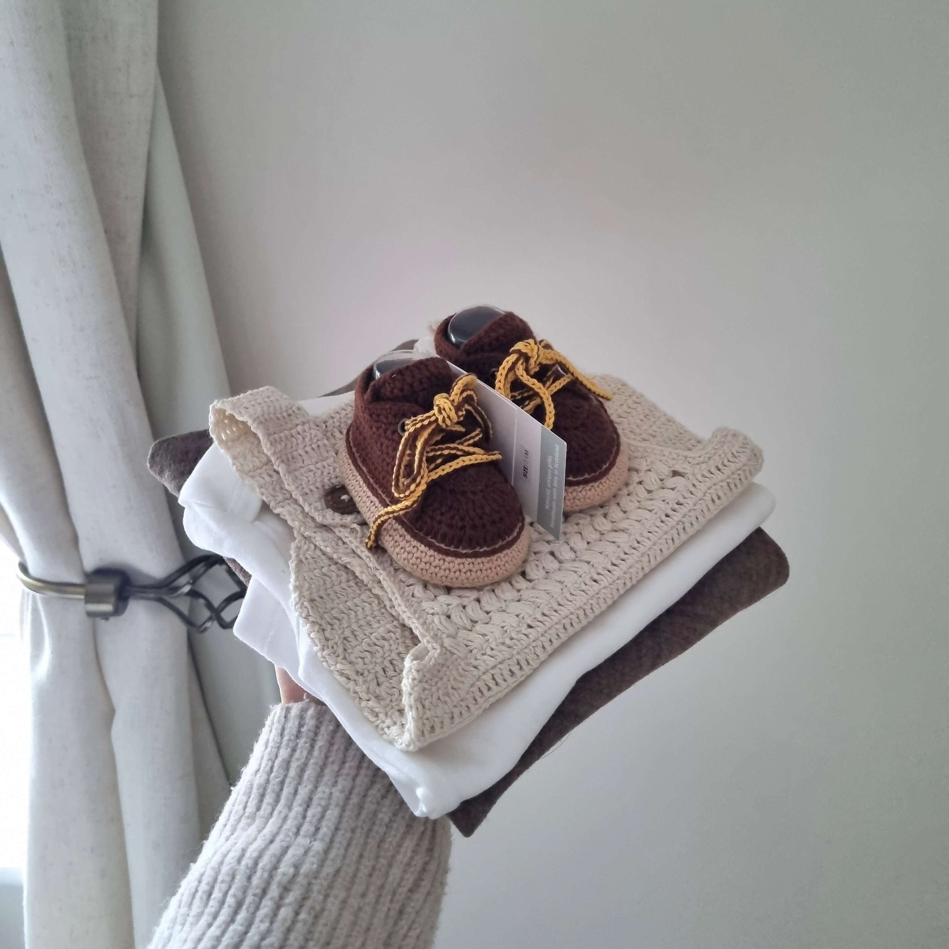 Hand-crocheted crochet baby boots in brown with lace-up design, displayed on top of folded clothing.