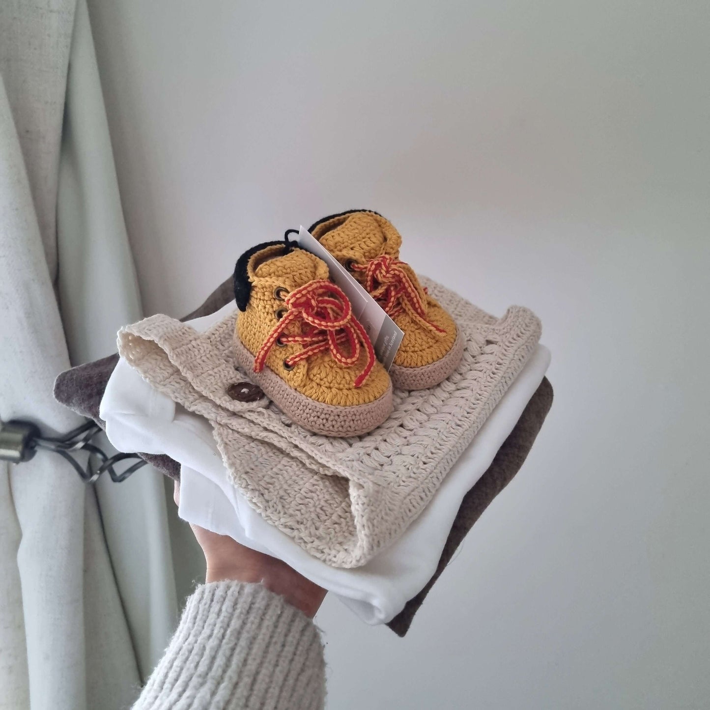 Hand-crocheted crochet baby work boots in yellow with orange laces resting on a stack of textile items.