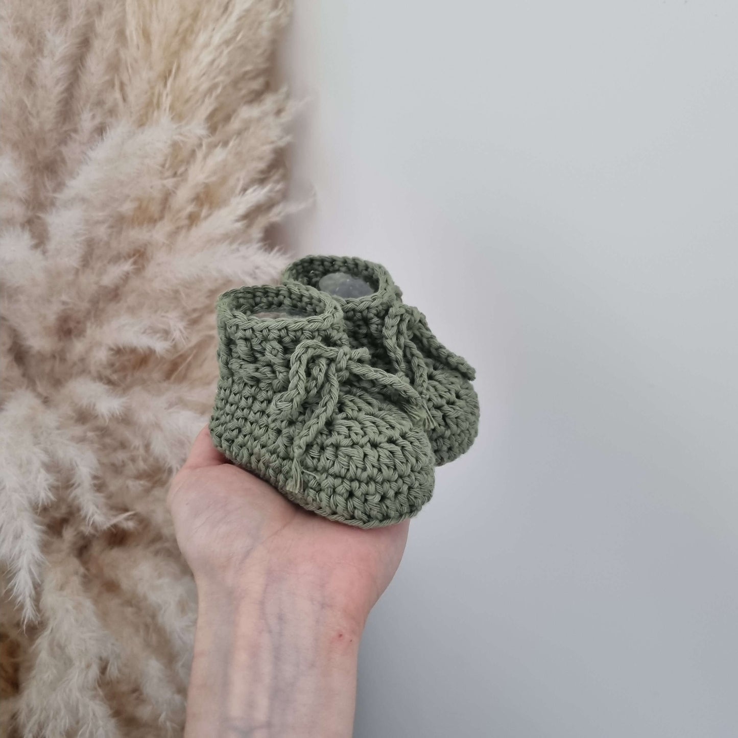 Green baby hat and boots set, featuring cozy crocheted boots with tie details held in a hand.