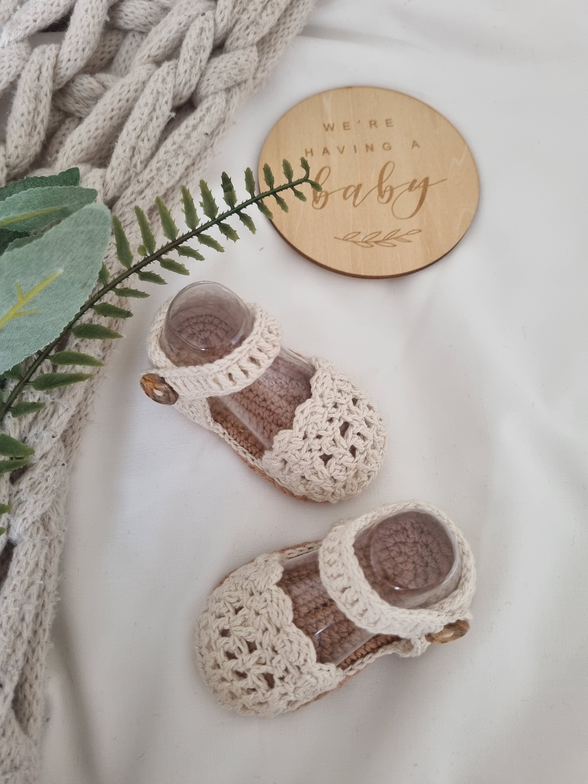 Pair of crocheted baby shoes with a wooden 'We're Having a Baby' sign on a soft surface.