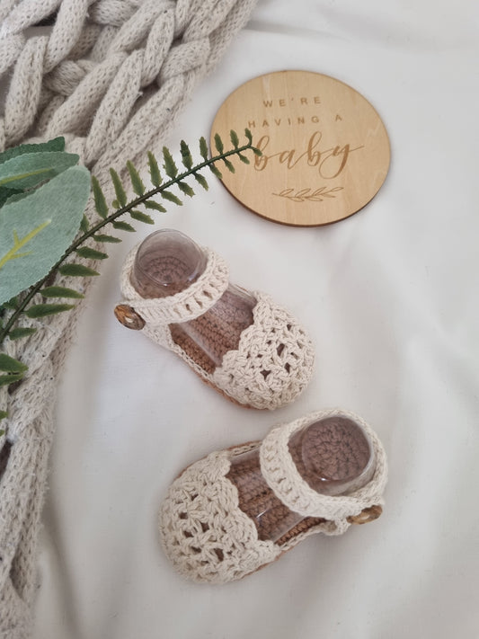 Pair of crocheted baby shoes with a wooden 'We're Having a Baby' sign on a soft surface.
