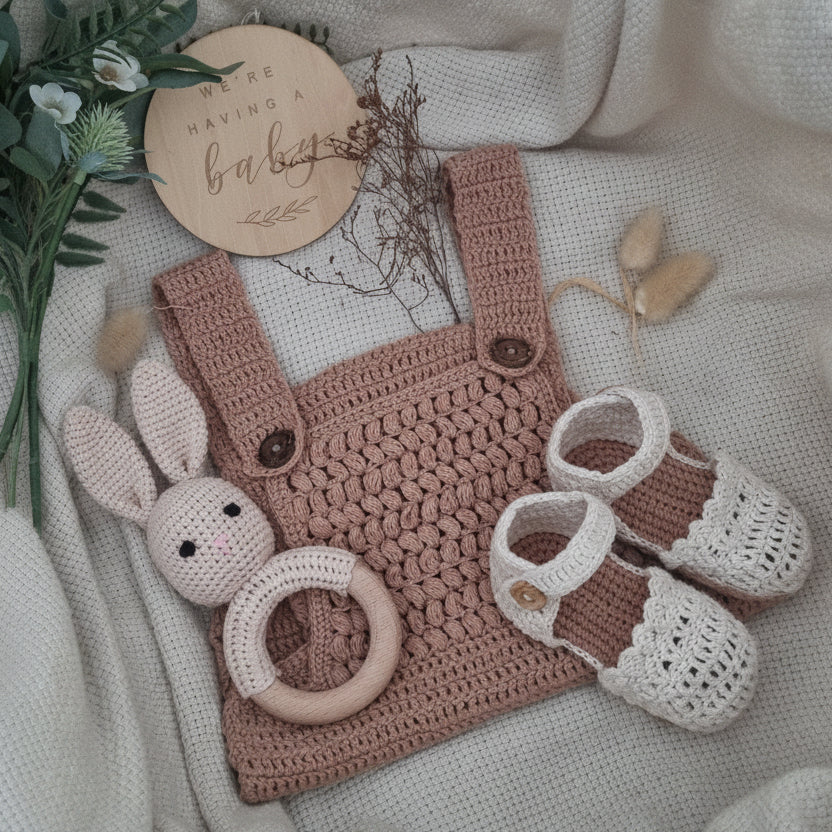 Crocheted baby outfit with overalls, shoes, and teething ring on a soft fabric background.