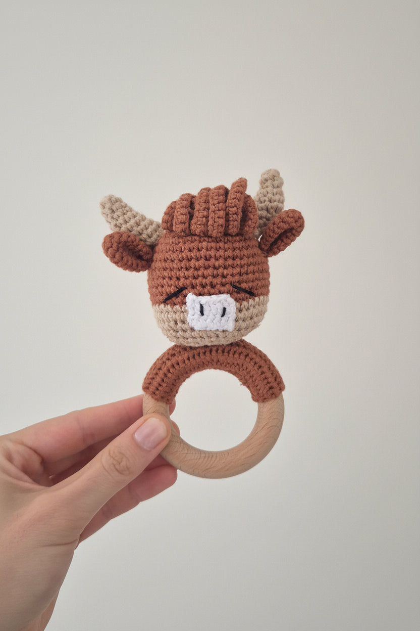 A hand holding a crocheted cow baby teether with a wooden ring handle against a plain light gray background.