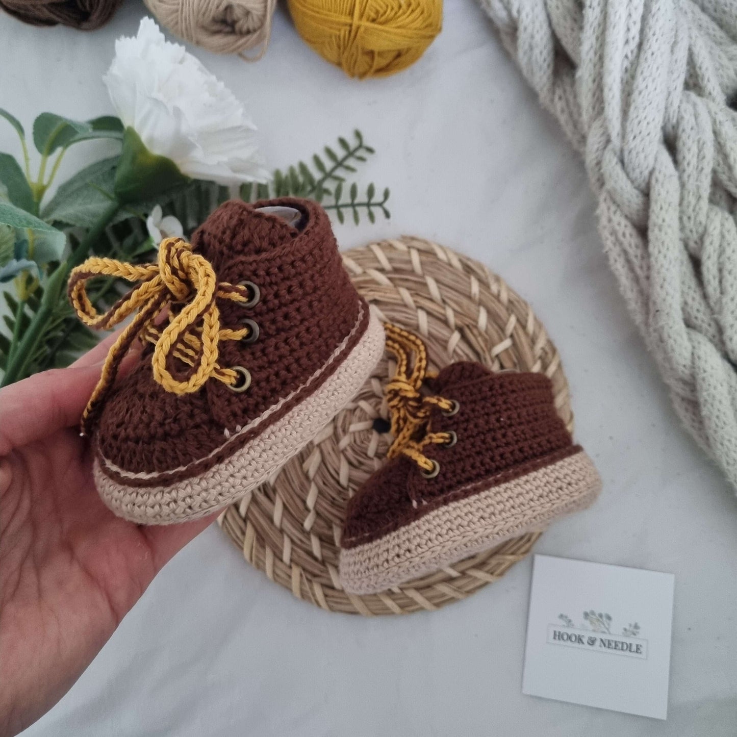Crochet baby boots in hand, featuring a lace-up design and soft, flexible structure on a decorative mat.