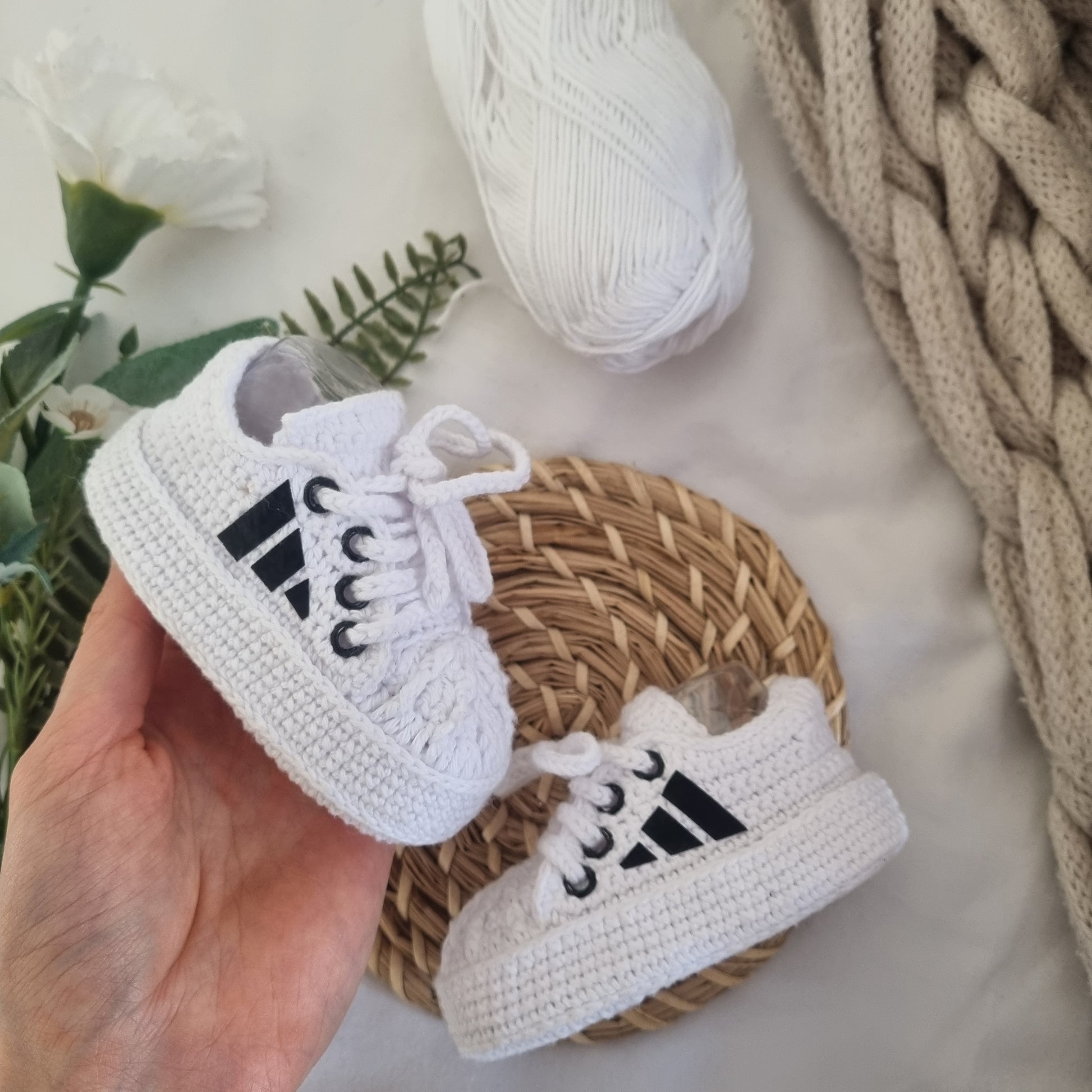 White baby sneakers with black stripes held by a hand, placed on a woven mat with a textured blanket and flowers in the background.