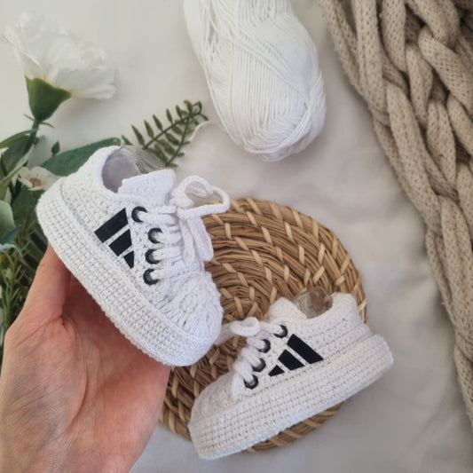 White baby sneakers with black stripes held by a hand, placed on a woven mat with a textured blanket and flowers in the background.
