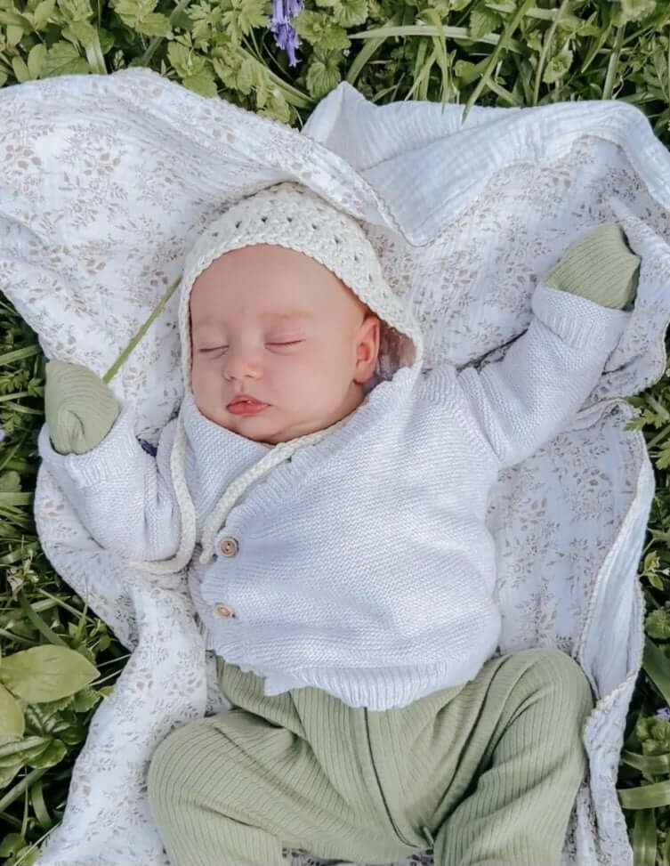 A baby in a bonnet and green pants is peacefully laying on the grass, enjoying a sunny day outdoors.