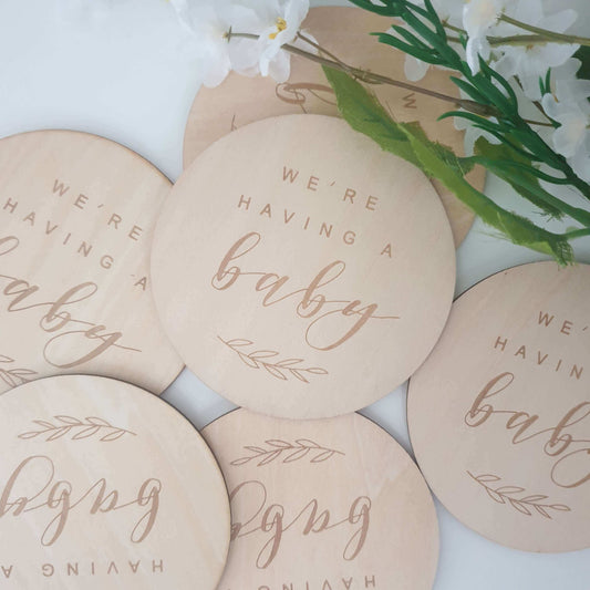 Wooden plaque with the text 'We're having a baby' in a circular shape, placed on a white surface with decorative flowers in the background.