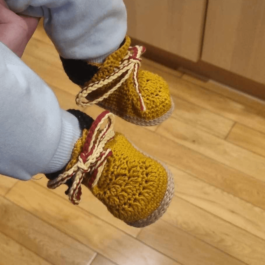 A person proudly holds their baby, showcasing a pair of Timberland style crocheted sneakers, displaying their unique and vibrant design.